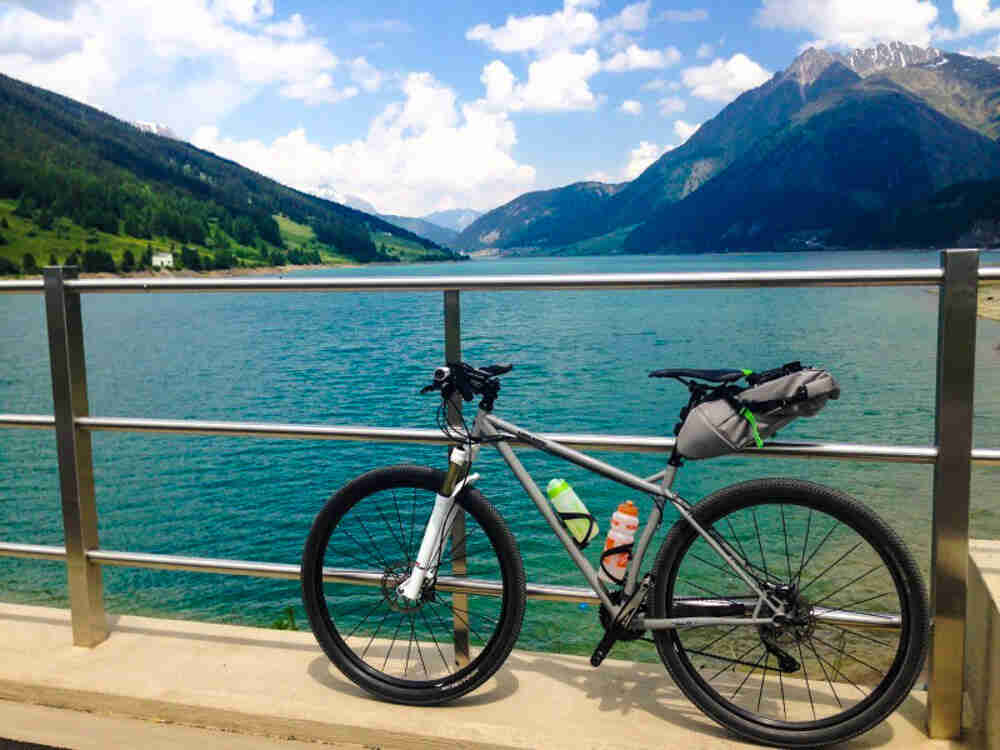 Left side view of a gray Surly bike, parked against a rail in front of a turquoise blue lake in the mountains