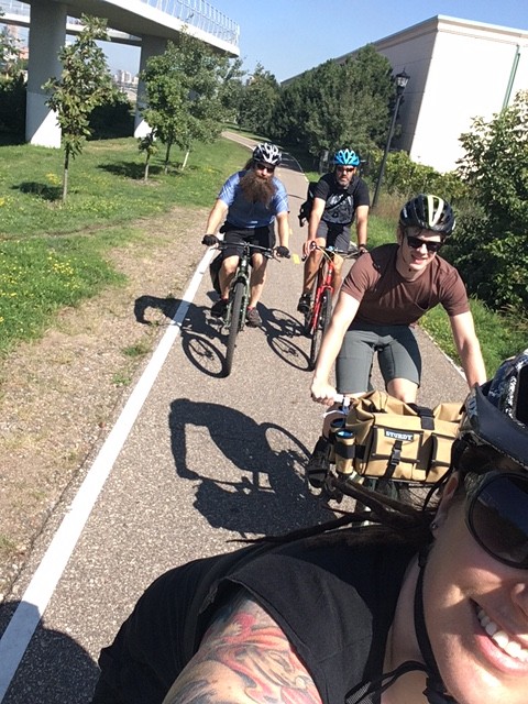 Front view of a group of cyclists riding their bike on a paved trail, with a building and a bridge in the background