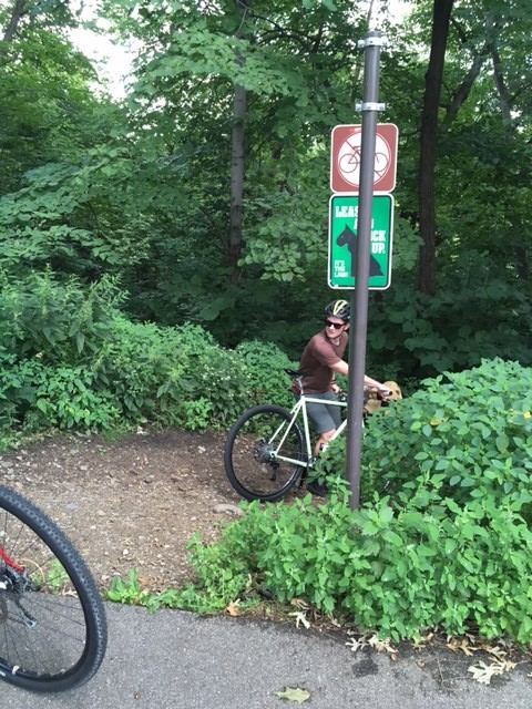 A cyclist looking back while walking into the woods with a bike, a the beginning of a trail