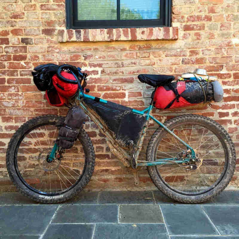 Left profile of a muddy green Surly bike, loaded with gear, parked on a sidewalk, in front of a red brick wall