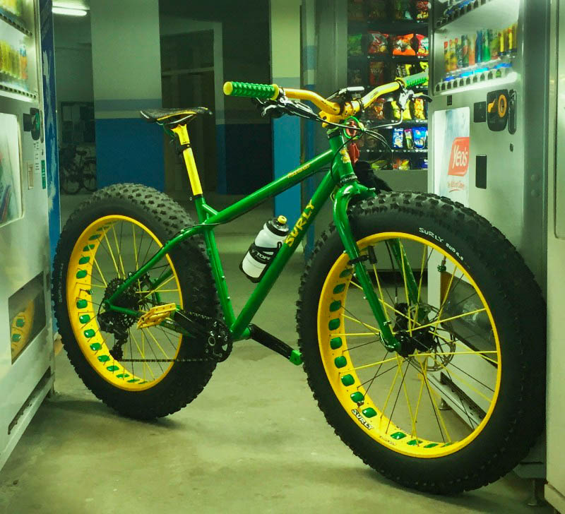 Front right, angled view of a green Surly fat bike, with yellow rims, wedged between two vending machines in a hallway