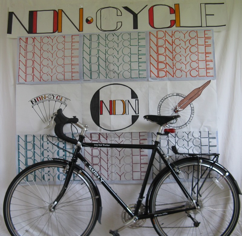 Left side view of a black Surly Long Haul Trucker bike, parked in front of wall with a banner showing NONCYCLE on it