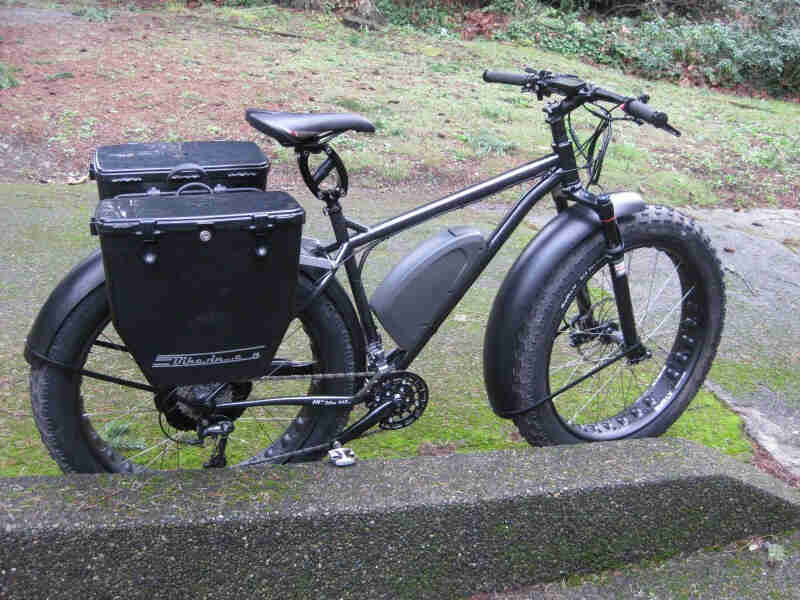Right side view of a black Surly fat bike with fenders and rear, hard side saddlebags, parked behind a short cement wall