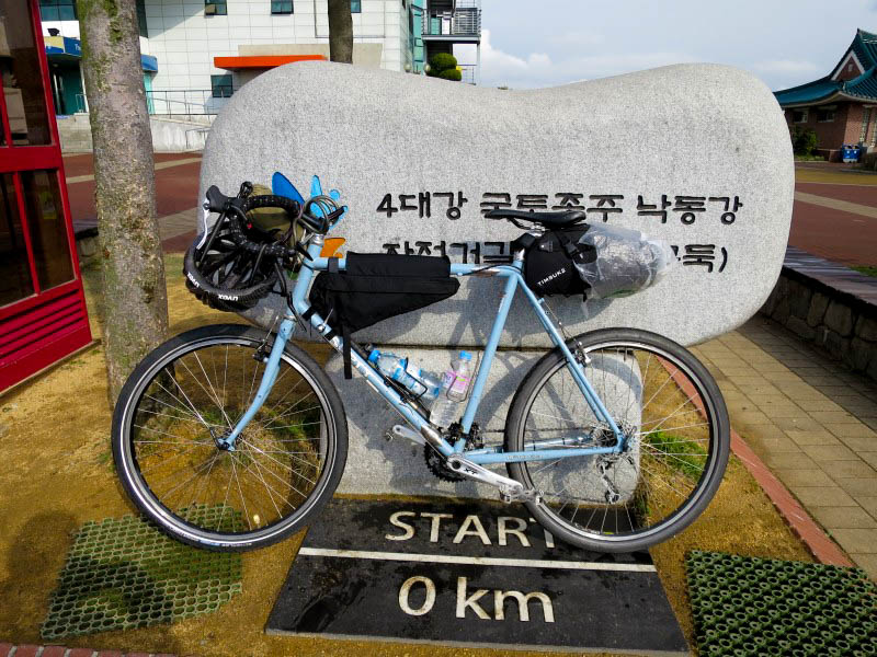 Left side view of a blue Surly bike in front of a rock sculpture, next to a sidewalk