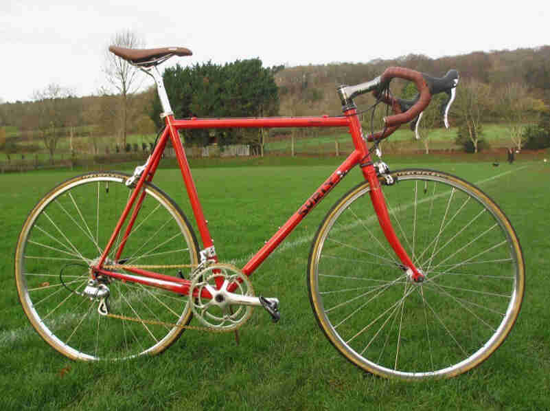 Right profile of a red Surly road bike, standing on a the side of a soccer field, with trees and hills in the background