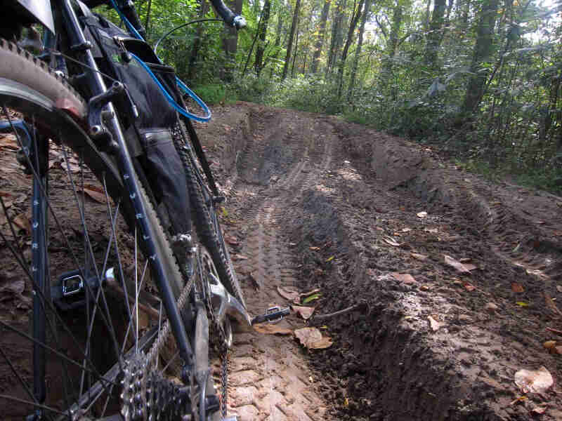 Zoom in of the right side of a black Surly bike, facing down a muddy trail in the woods