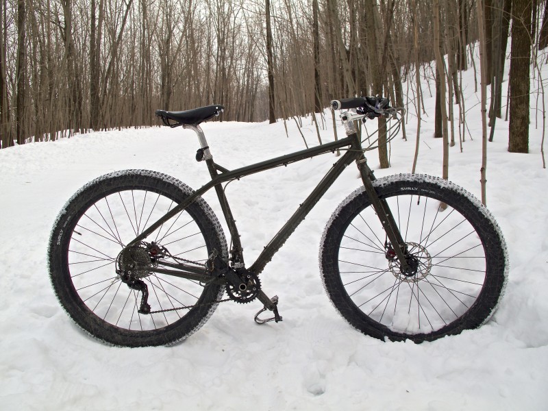 Right side view of a black Surly bike, standing in a snow covered clearing in the woods, with bare trees