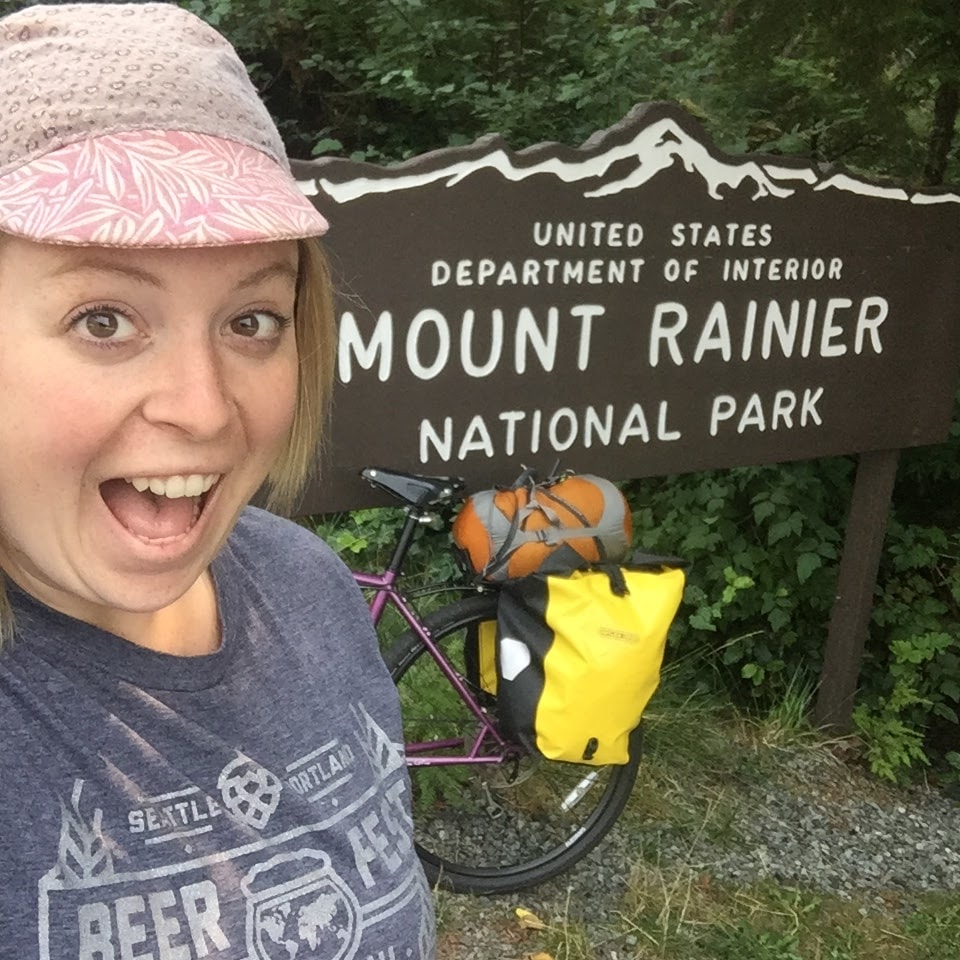 Excited cyclist takes a selfie in front of a Mount Rainer National Park sign and a bike loaded with gear