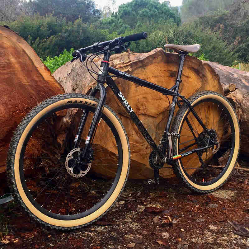 Front left angled view of a Surly Karate Monkey bike, black, leaning against 2 boulders, with trees in the background