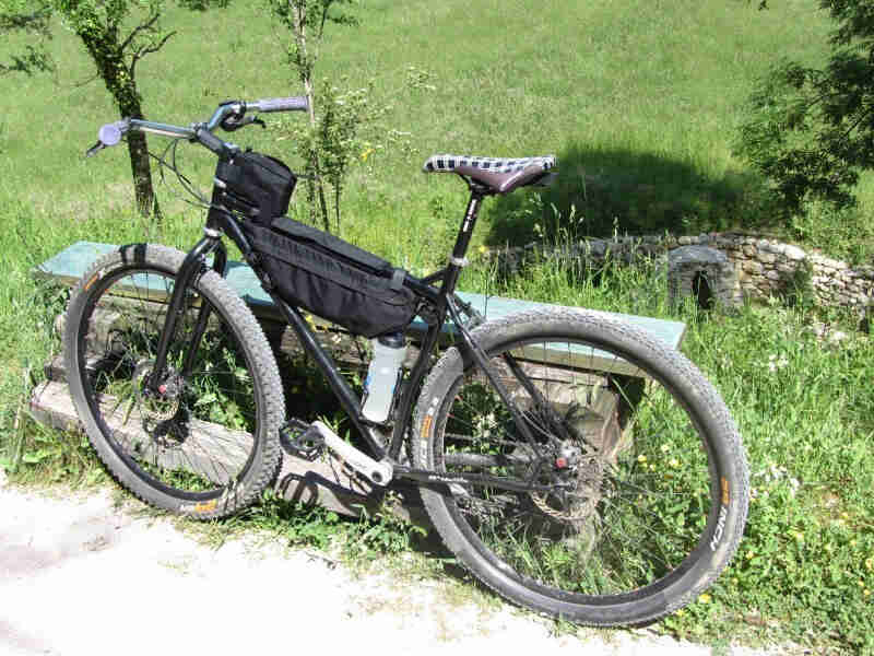 Right side view of a black Surly bike, parked along a wood bench next to a dirt trail, with a grass field in background