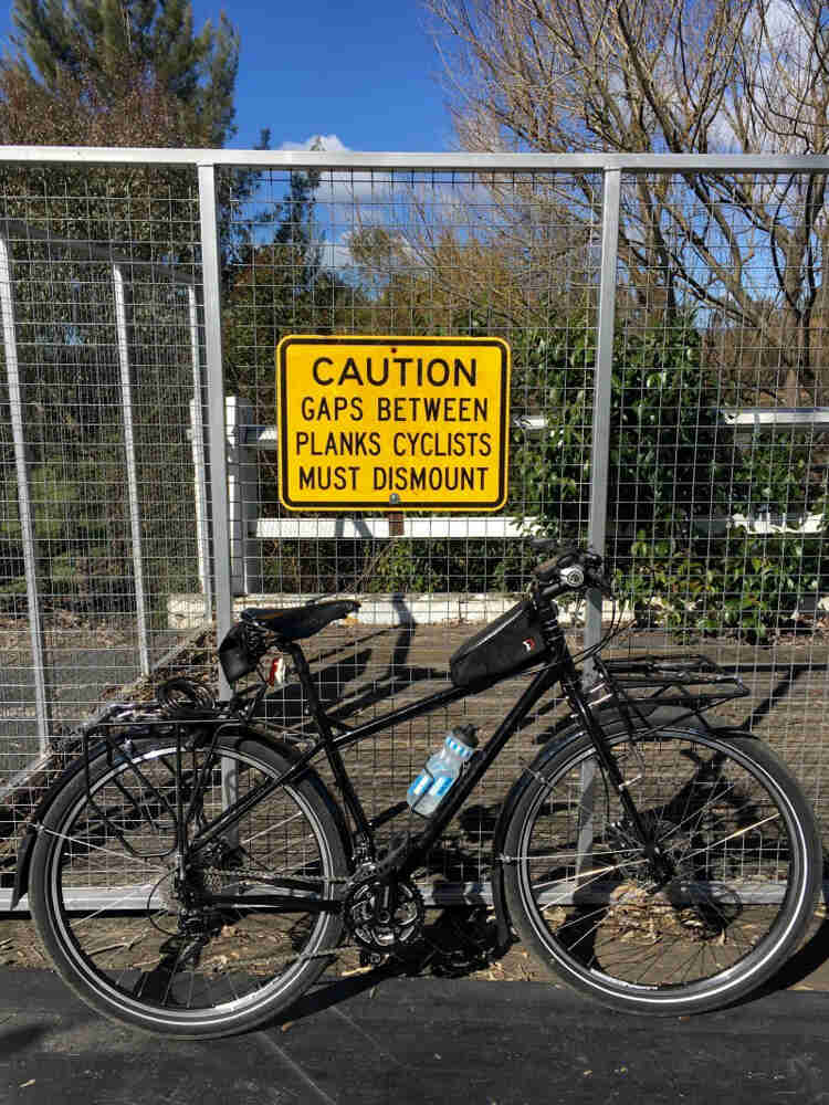 Right profile of a Surly bike, black, parked on pavement, against a steel mesh cage with a caution sign on it