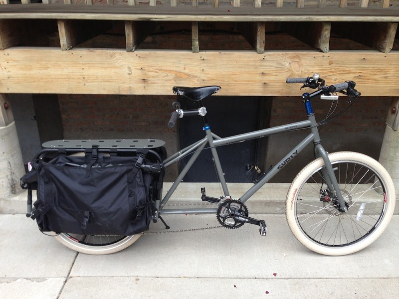 Right side view of a gray Surly Big Dummy bike with white tires, parked on a sidewalk, beside a wood deck