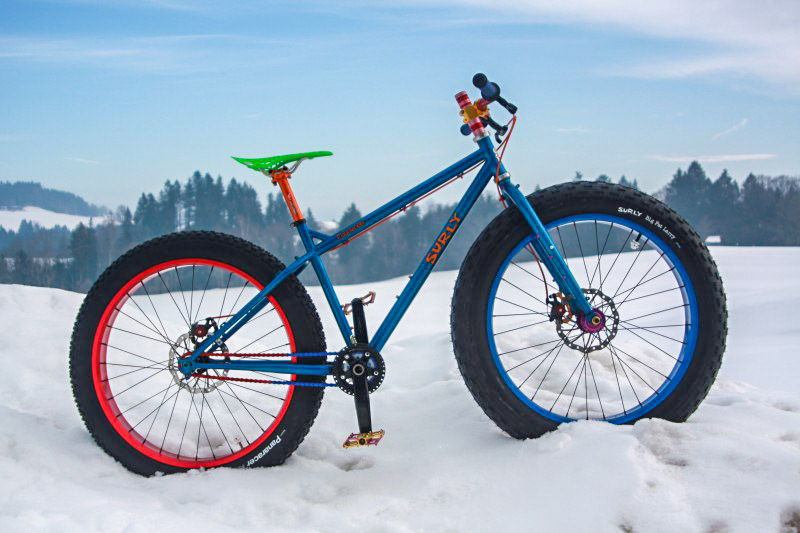 Right profile of Surly fat bike, blue, parked in a snow covered field, with pine trees in the background