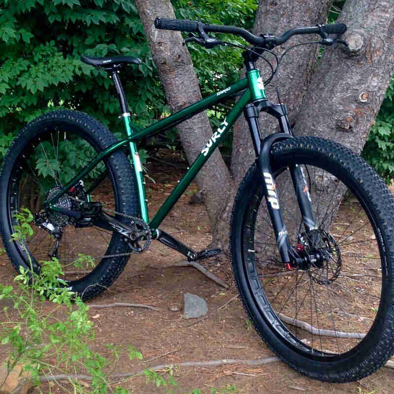 Front, right side view of a green Surly Krampus bike, parked at the base of a tree with 3 trunks, and bushes behind