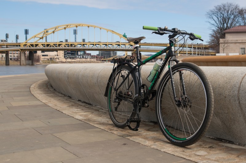 Front, right side view of a black Surly bike, leaning on a cement, boardwalk barrier, with a bridge in the background
