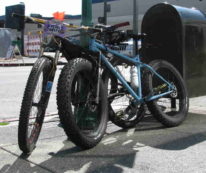 Front, right side view of two Surly bikes, leaning on a light pole, on a sidewalk with city buildings across the street