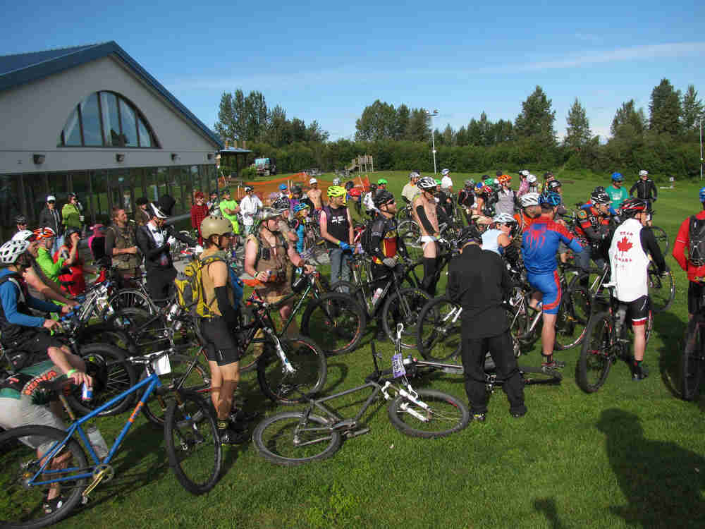 A group of cyclists and their bikes, gathered together on a grass field, outside of a building on the left