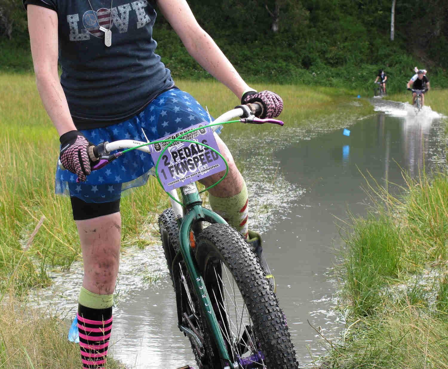 Front view of a cyclist, standing over a green Surly Krampus bike, in a flooded grass field with trees in the background