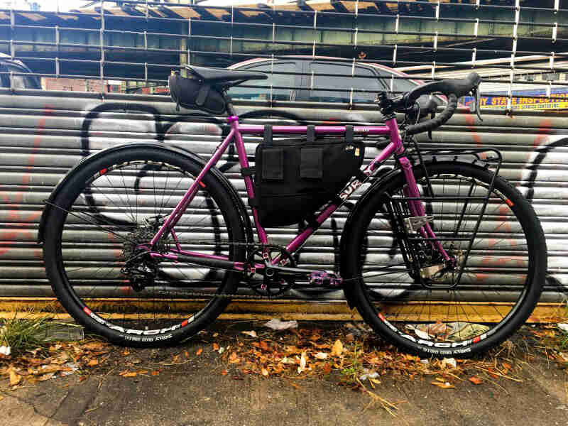 Right side view of a Surly bike, purple, on a sidewalk and leaning against steel fence