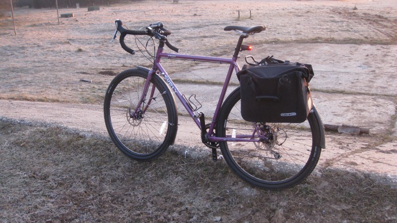 Right side view of a purple Surly bike with rear bags, parked beside a dirt trail, with a field in the background