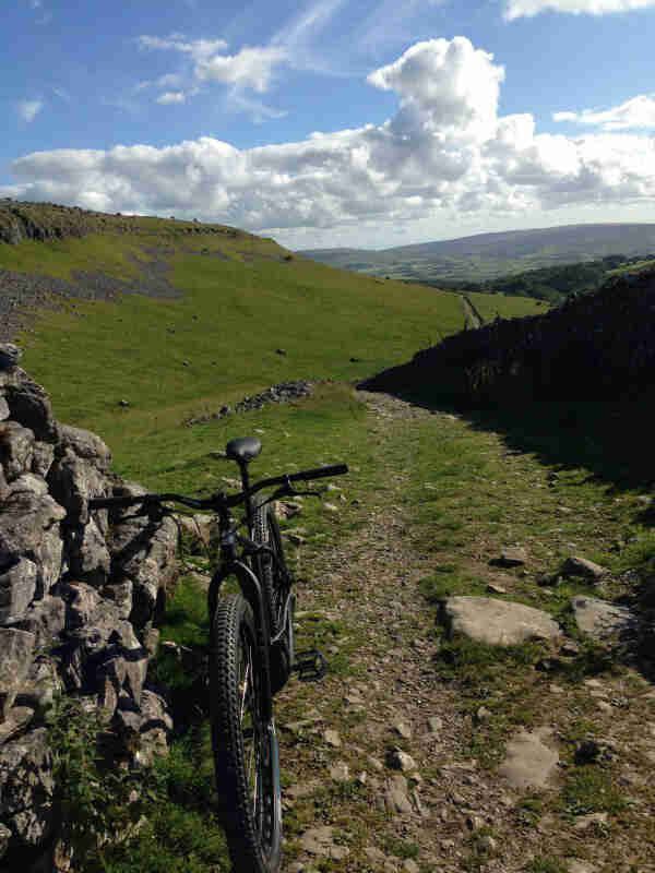 Front view of a Surly Krampus bike, black, facing upward on a gravel trail, with green hills in the background