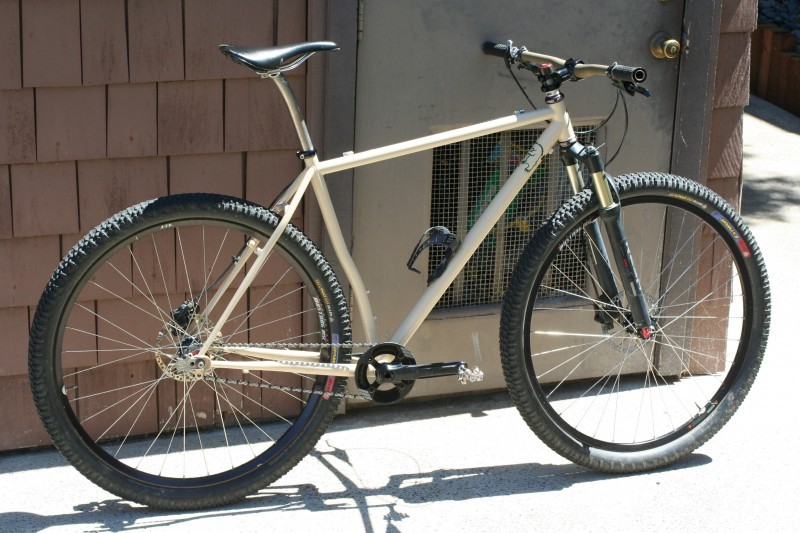 Right side view of a tan Surly bike, parked on a sidewalk, leaning against a door on a building with brown wood siding