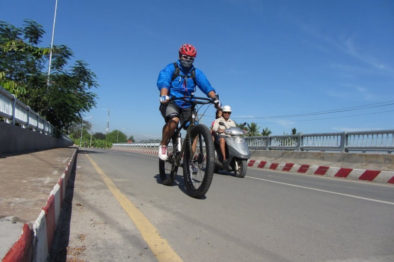 Front, right side view of a cyclist, riding a Surly bike across a paved bridge, with a motor scooter next to them