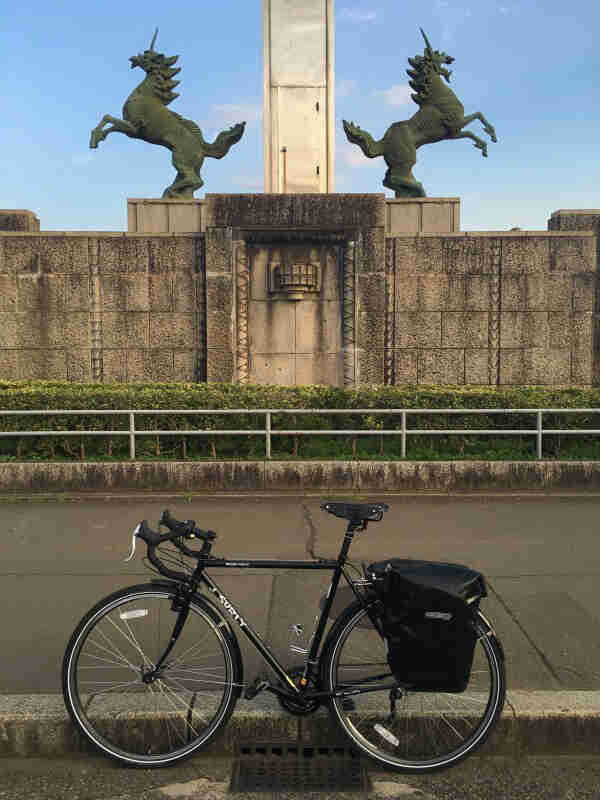 Left profile of a Surly bike, parked on a street curb, with a stone platform with unicorns on top, in the background