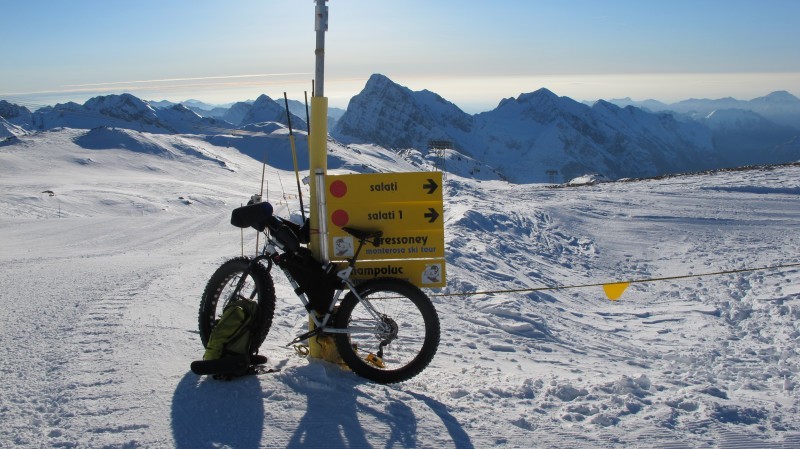 Left side view of a white Surly fat bike with gear, leaning against a pole, in a snow covered field in the mountains