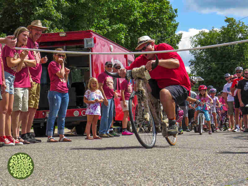Front view of a cyclist riding a bike with bull horn handlebars towards a limbo bar, with spectators watching