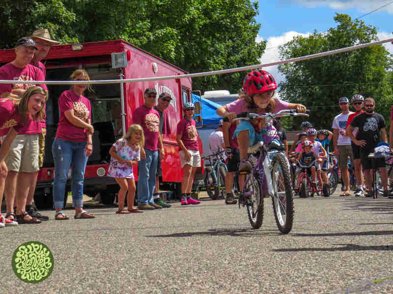 Front view of a child on a bike, riding under a limbo bar, with spectators watching