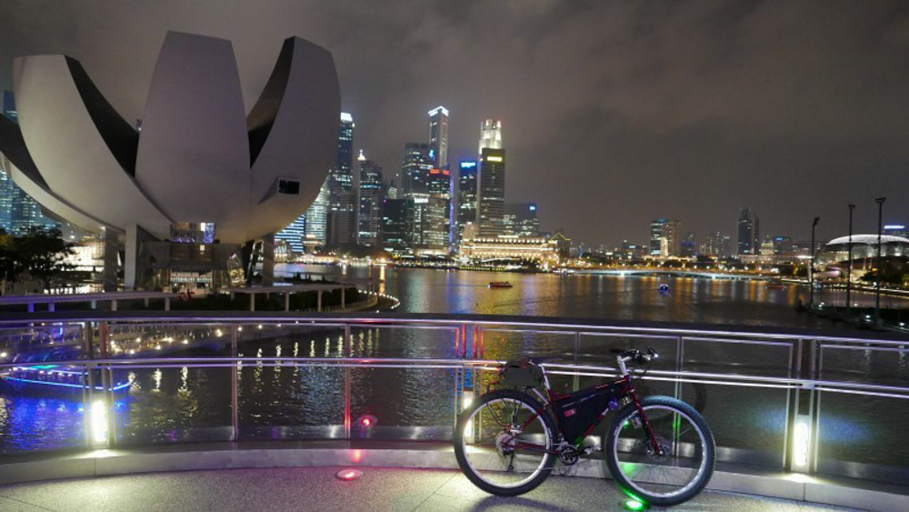 Right side view of a bike loaded with gear, parked on a platform over a bay, with a city in the background at night