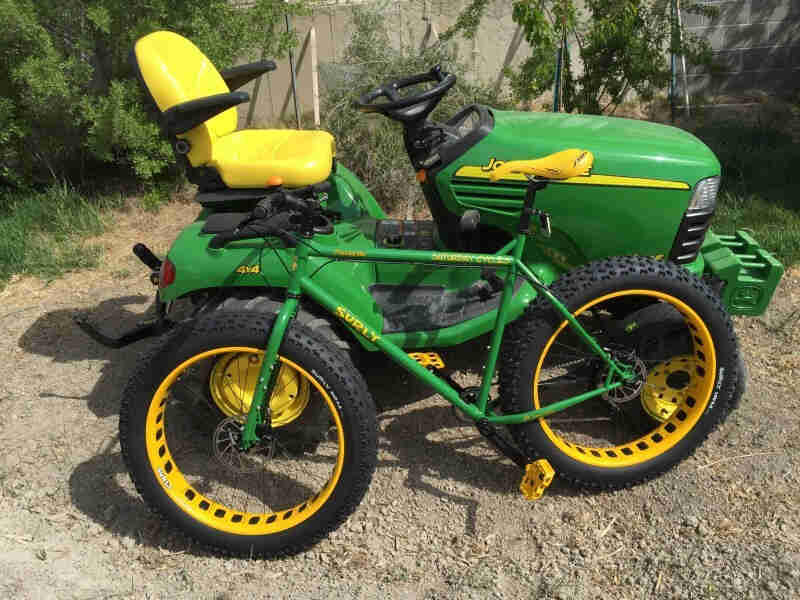 Left side view of a green and yellow Surly fat bike, parked on gravel alongside a John Deere yard tractor