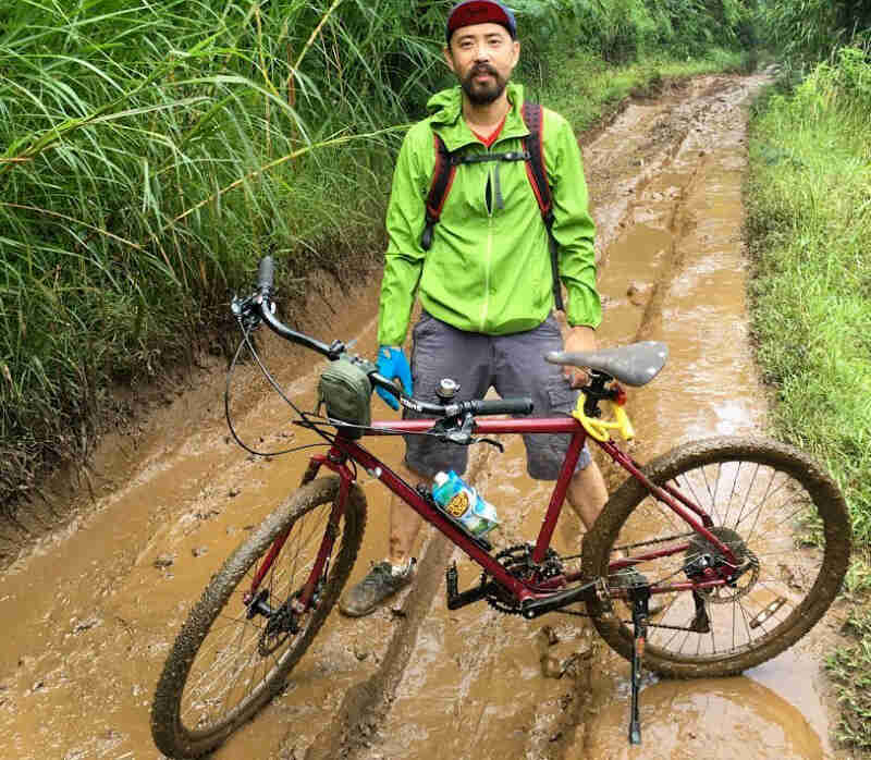 Front view of a cyclist standing in mud behind a bike parked across a muddy jungle road