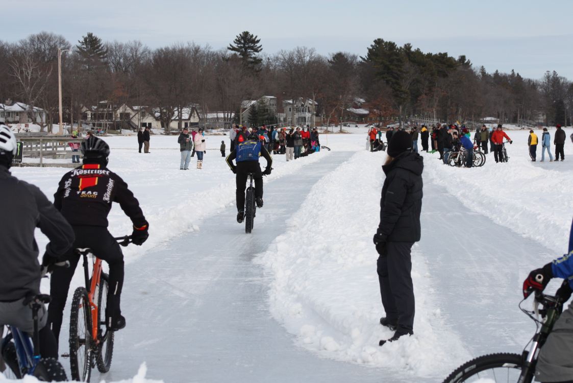 Rear view of cyclists riding their bike downs a straight-away icy path on a frozen, snow covered lake, with spectators