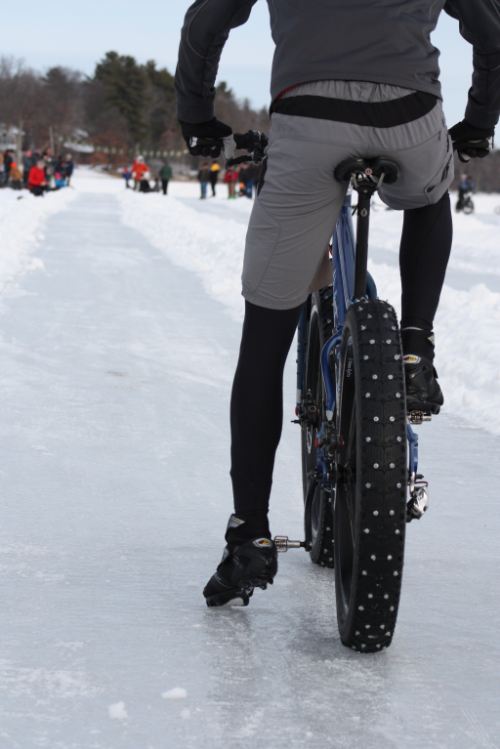 Rear view of a cyclist sitting on a blue Surly fat bike with studded tires, facing down a path on a snowy, frozen lake