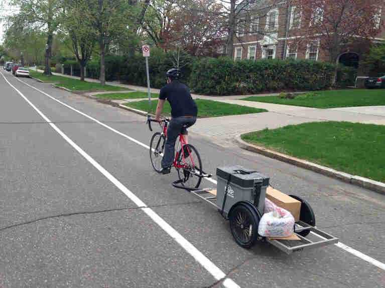 Rear, left side view of a cyclist riding a bike with trailer, in the bike lane on a street, with houses to the right