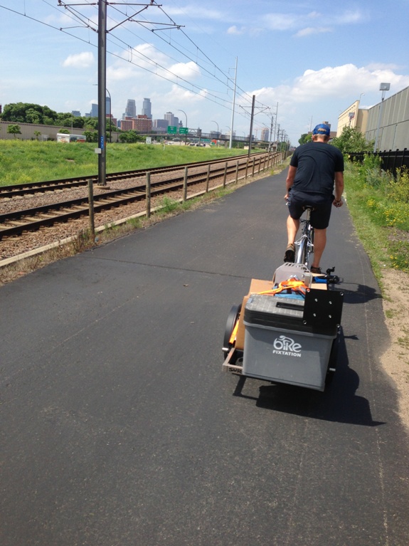 Rear view of a cyclist riding a bike with trailer, on a paved trail along light rail tracks, with a city skyline ahead