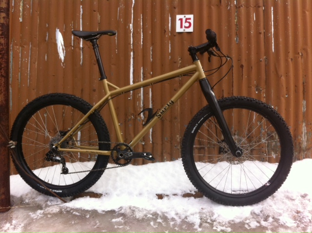 Right side view of a mustard yellow Surly Instigator bike, parked on snow, leaning on a corrugated steel wall