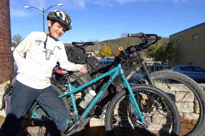 Front view of a young cyclist, leaning on the seat of a turquoise Surly troll bike, in front of an olive Surly fat bike