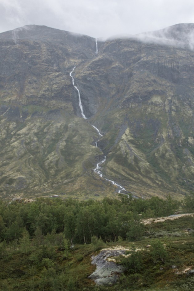 A mountain with cloudy peak and a narrow stream flowing done to the base
