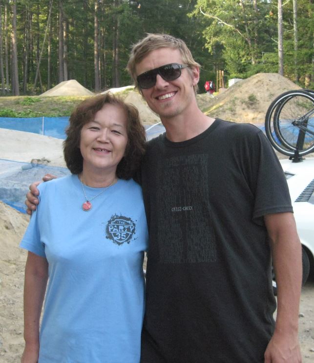 Front view of two people posing together, with a BMX track and a forest in the background