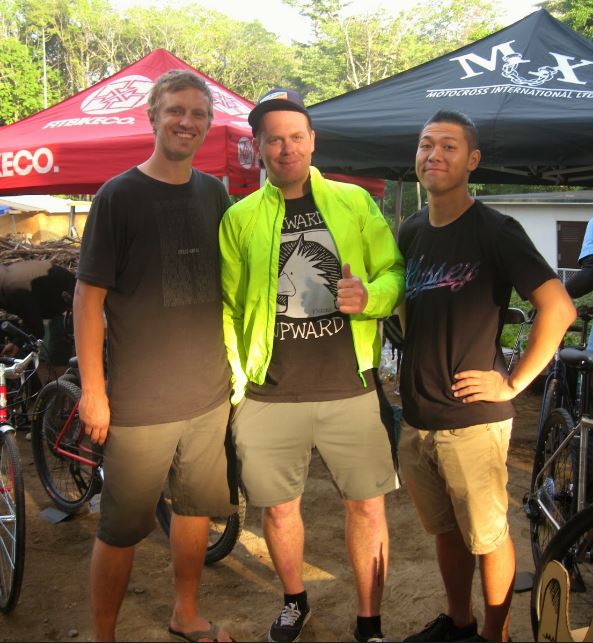 Front view of three people, shoulder to shoulder, posing for a picture with bikes, canopies and trees behind them