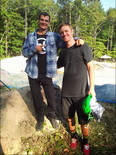 Front view of a person posing with BMX legend, Matt Hoffman, in front of a BMX track with a green forest behind it
