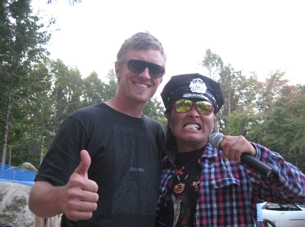 Front view of a person, posing next to a person wearing a police hat and mirrored sunglasses, with trees behind them