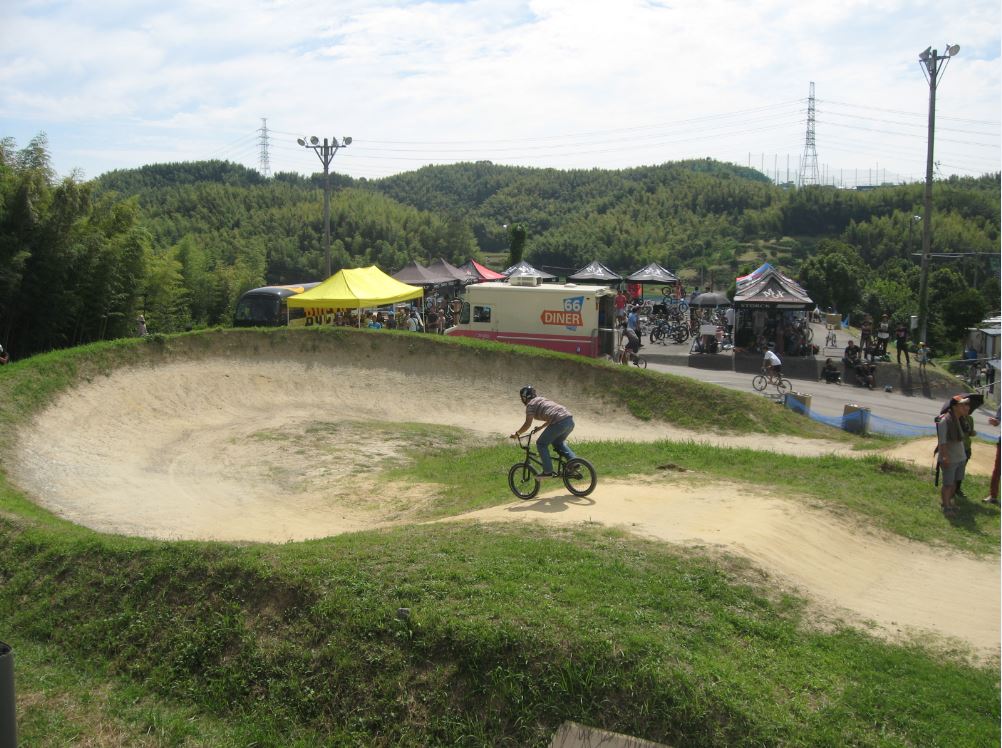 Right side view of a cyclist, riding over a mound on a dirt BMX track, with canopies and tree covered hills behind it