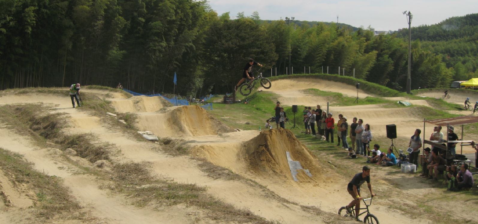 Right side view of a cyclist, high in the air, after jumping a bike off a ramp on a dirt BMX track with people watching