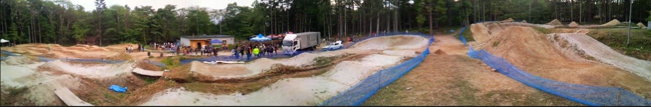 Panoramic view of a dirt BMX track with people standing around, with a pine forest behind it