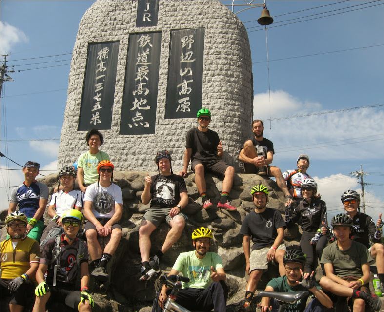 Front view of a group of cyclists posing, sitting around the stone base of a brick monument with Japanese text on it