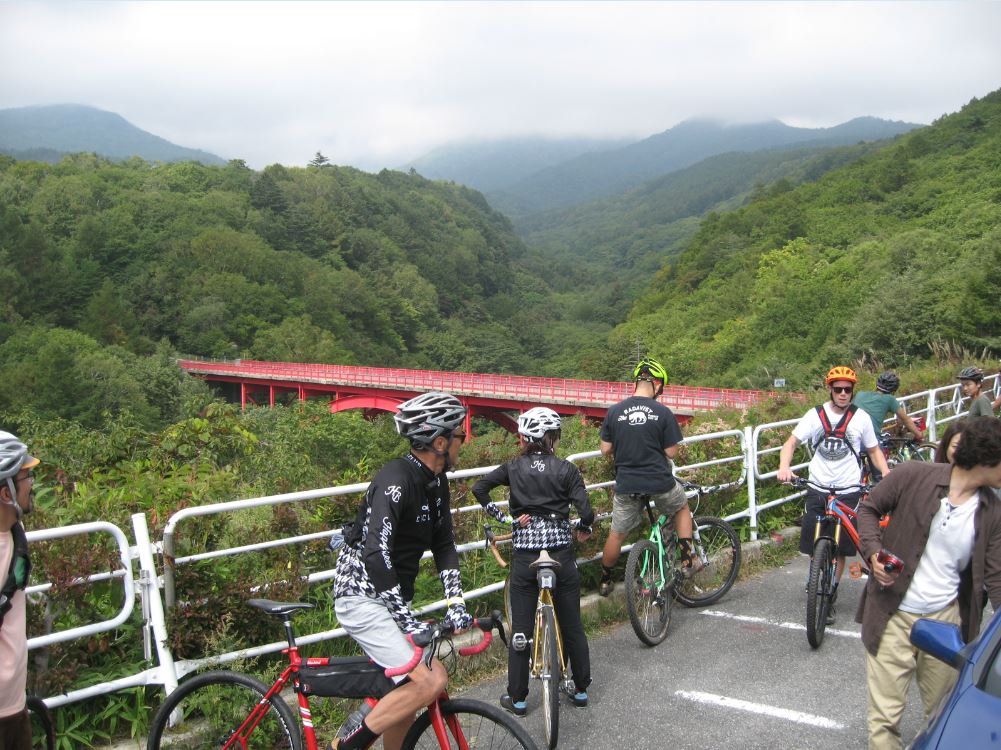 Cyclists standing with their bikes on a paved trail in the tree covered mountains, with a bridge in a valley, behind